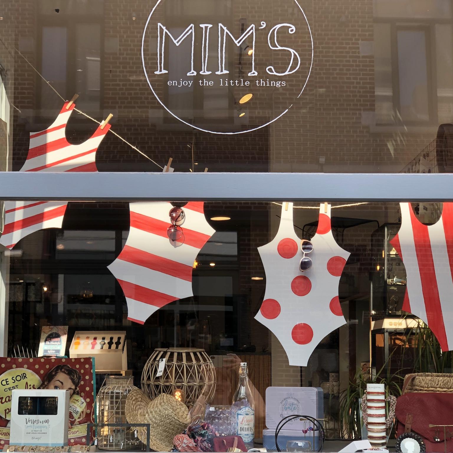 Vitrine de la boutique Mim's à Leuze-en-Hainaut avec maillots de bain une pièce blanc et rouge suspendus sur un fil à linge, lunettes de soleil, montres, chapeaux et décoration estivale