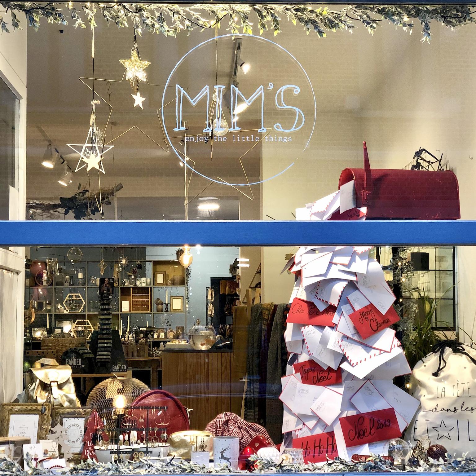 Vitrine de Noël de la boutique Mim's à Leuze-en-Hainaut avec une boîte aux lettres rouge débordant de cartes postales rouges et blanches, entourée de bijoux et objets de décoration rouges et dorés